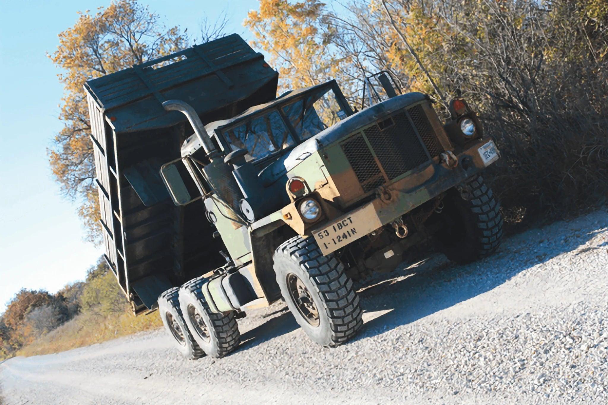 A PIERCE Heavy Duty Flat Bed Dump Kit is installed on a military dump truck with its bed raised, parked on a gravel road surrounded by trees with autumn foliage.