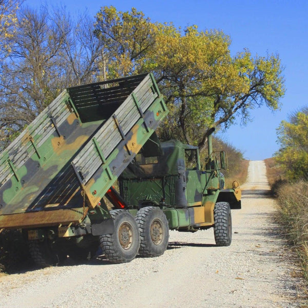 A military dump truck equipped with a PIERCE Heavy Duty Flat Bed Dump Kit is parked on a rural, tree-lined gravel road with its bed raised.