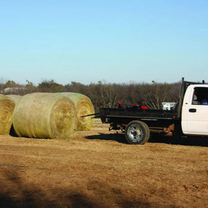 A PIERCE Truck Bed Gooseneck Hay Bale Mover is mounted on a truck, transporting a large hay bale in a field with additional bales and trees in the background.