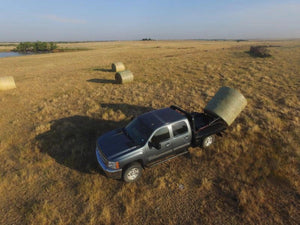 A black pickup truck equipped with a PIERCE Bolt-in Hydraulic Truck Bed Hay Bale Spear unloads a large hay bale in a field with more hay bales scattered nearby.