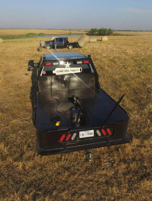 A black flatbed truck with COME AND TAKE IT text, parked beside hay bales and another truck in a dry field, is equipped with the PIERCE Truck Bed Gooseneck Hay Bale Mover for efficient bale handling.