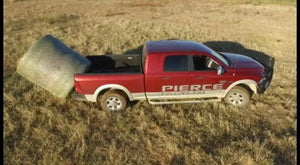 A red pickup truck in a field carries a large hay bale in its bed with the PIERCE logo, equipped with the PIERCE Truck Bed Gooseneck Hay Bale Mover.