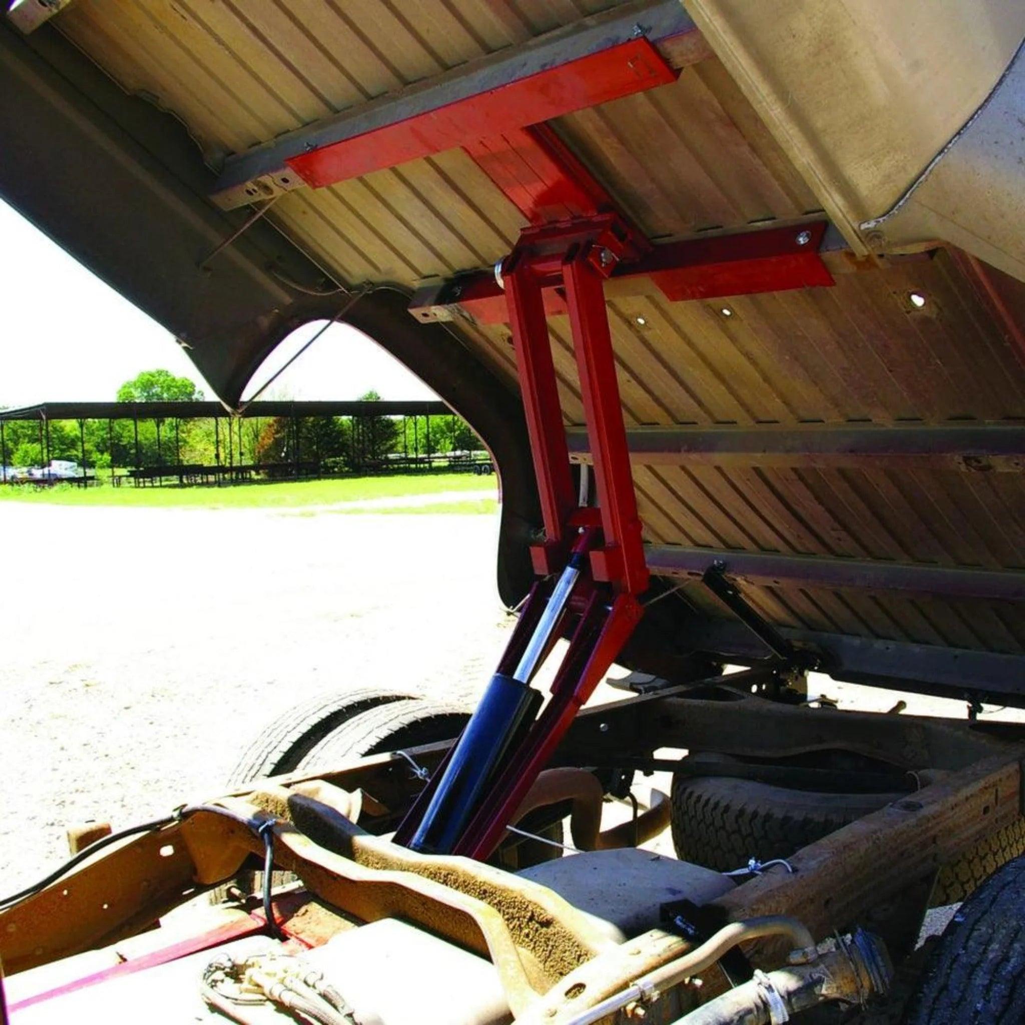 The Factory Bed Dump Kit by PIERCE is displayed under a truck's tilted cargo bed in a sunny outdoor setting, emphasizing its impressive lifting strength.