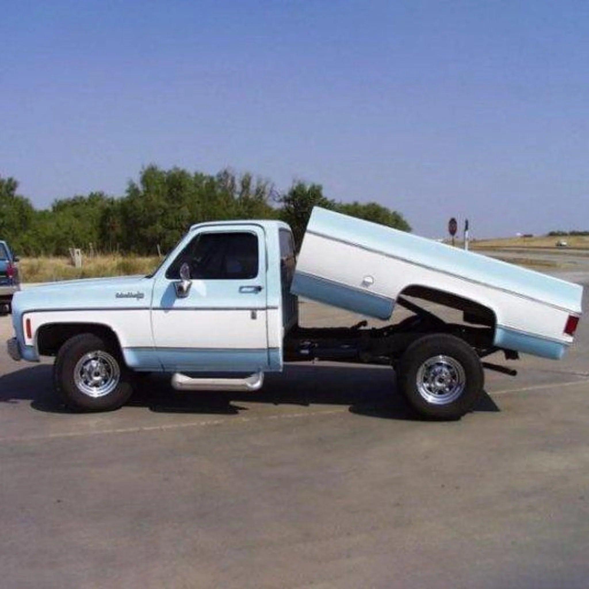 A pickup truck fitted with the PIERCE Factory Bed Dump Kit has its bed tilted upwards, revealing the frame and wheels underneath while resting on a paved road.
