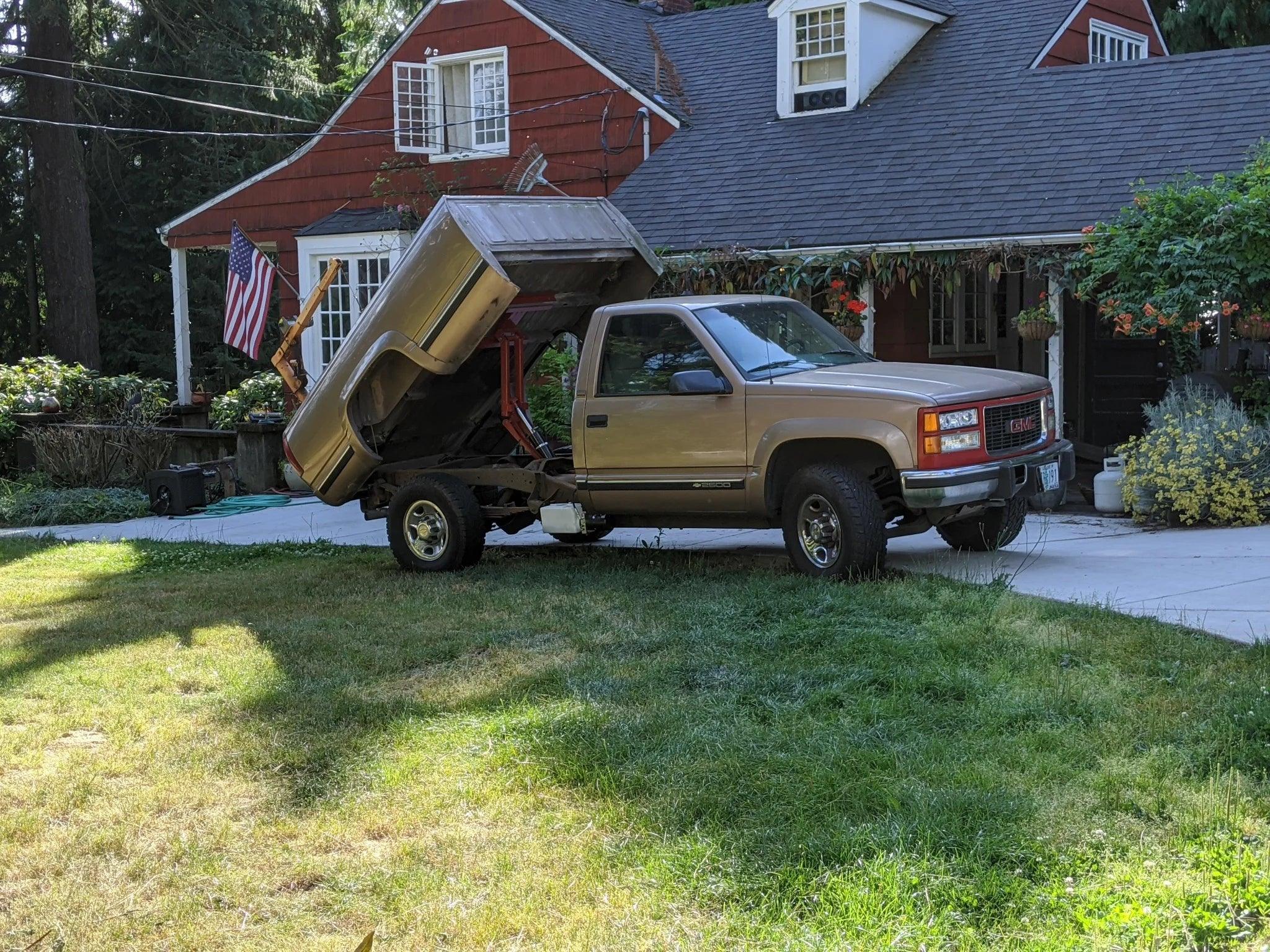 A pickup truck equipped with a PIERCE Factory Bed Dump Kit and raised bed is parked in front of a red house adorned with an American flag.
