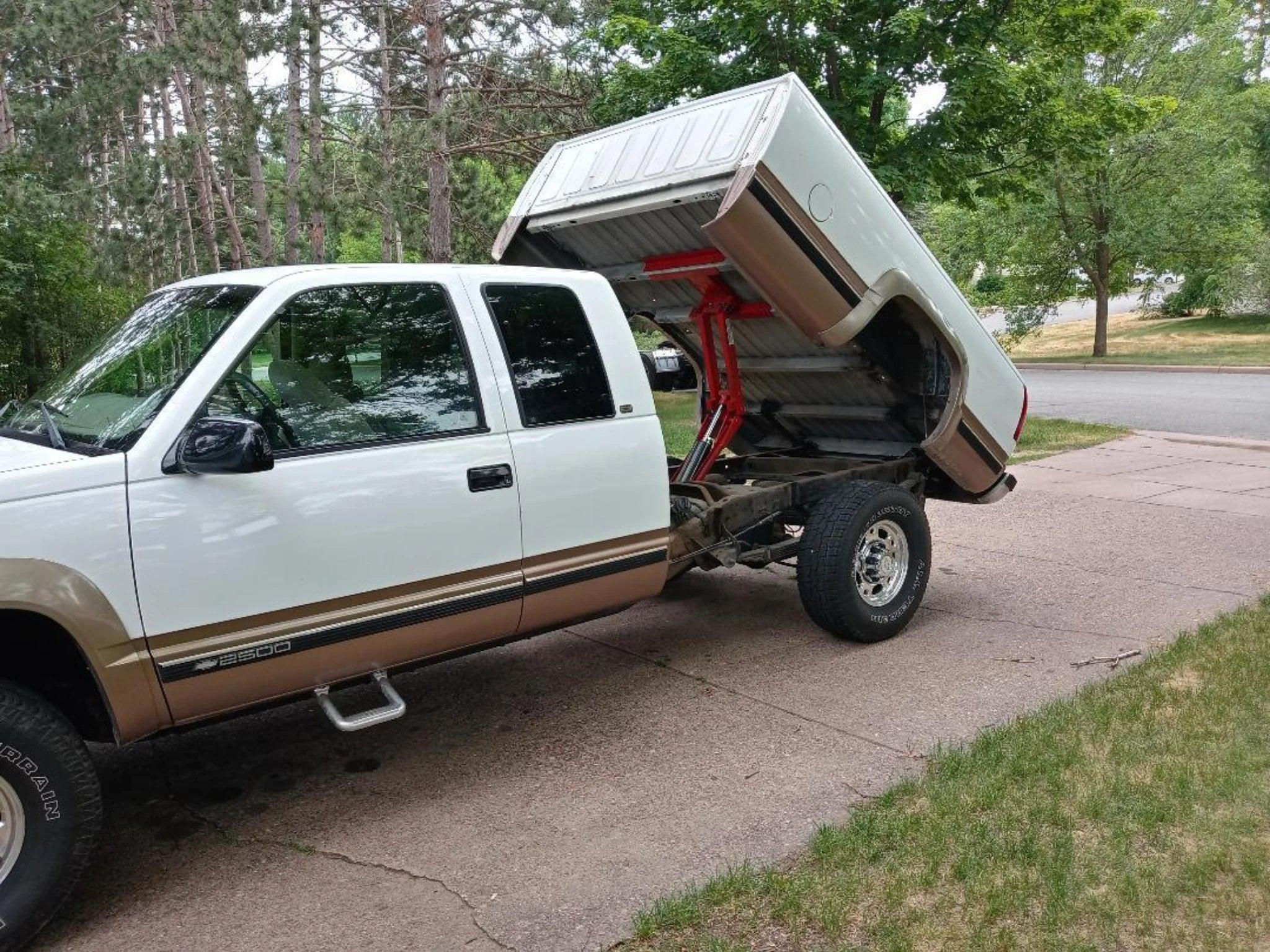A pickup truck equipped with the PIERCE Factory Bed Dump Kit, showcasing its robust lifting ability with the flatbed raised to reveal the frame, is parked on a driveway surrounded by greenery and trees.