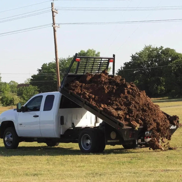 A white truck equipped with the PIERCE Light Duty Flat Bed Dump Kit unloads dirt onto a grassy field under a clear sky.
