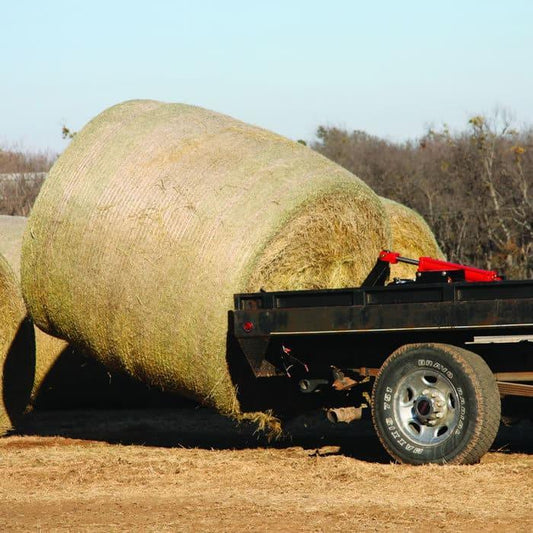 Large round hay bales rest beside a flatbed trailer with trees behind, ready to be loaded using the PIERCE Truck Bed Gooseneck Hay Bale Mover.
