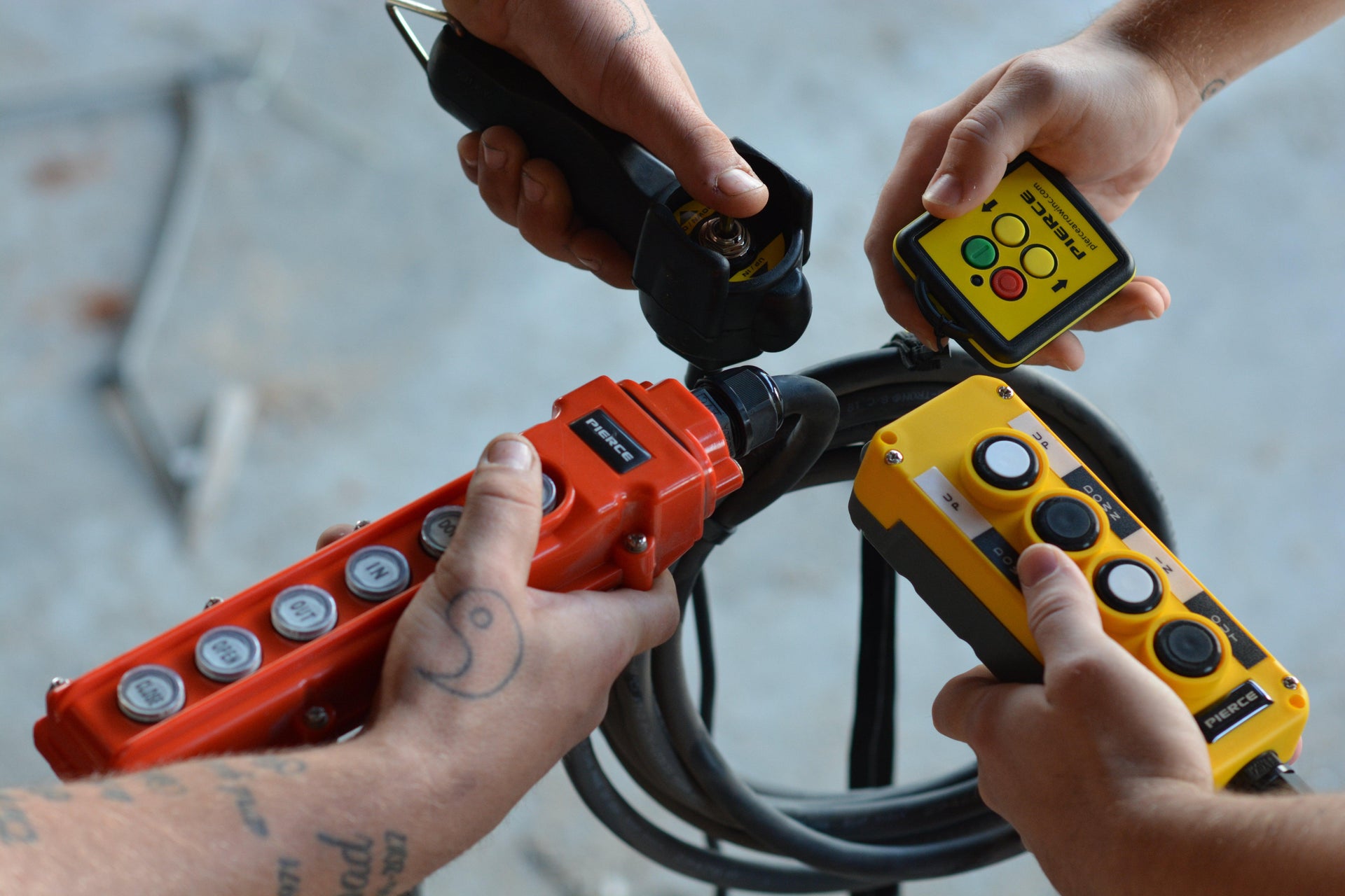 Four hands hold industrial remote controls and cables, operating heavy machinery or equipment.