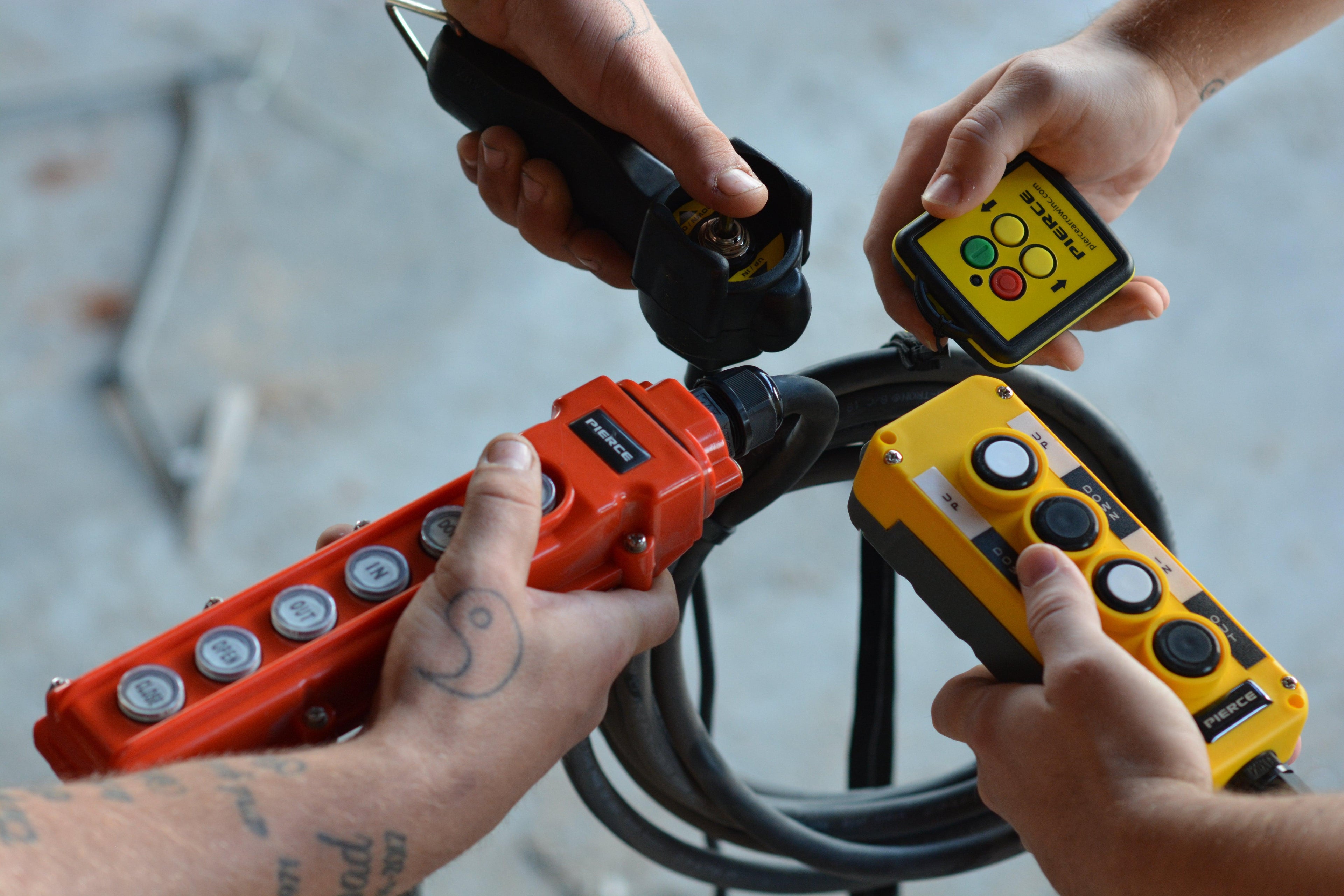 Four hands hold industrial remote controls and cables, operating heavy machinery or equipment.