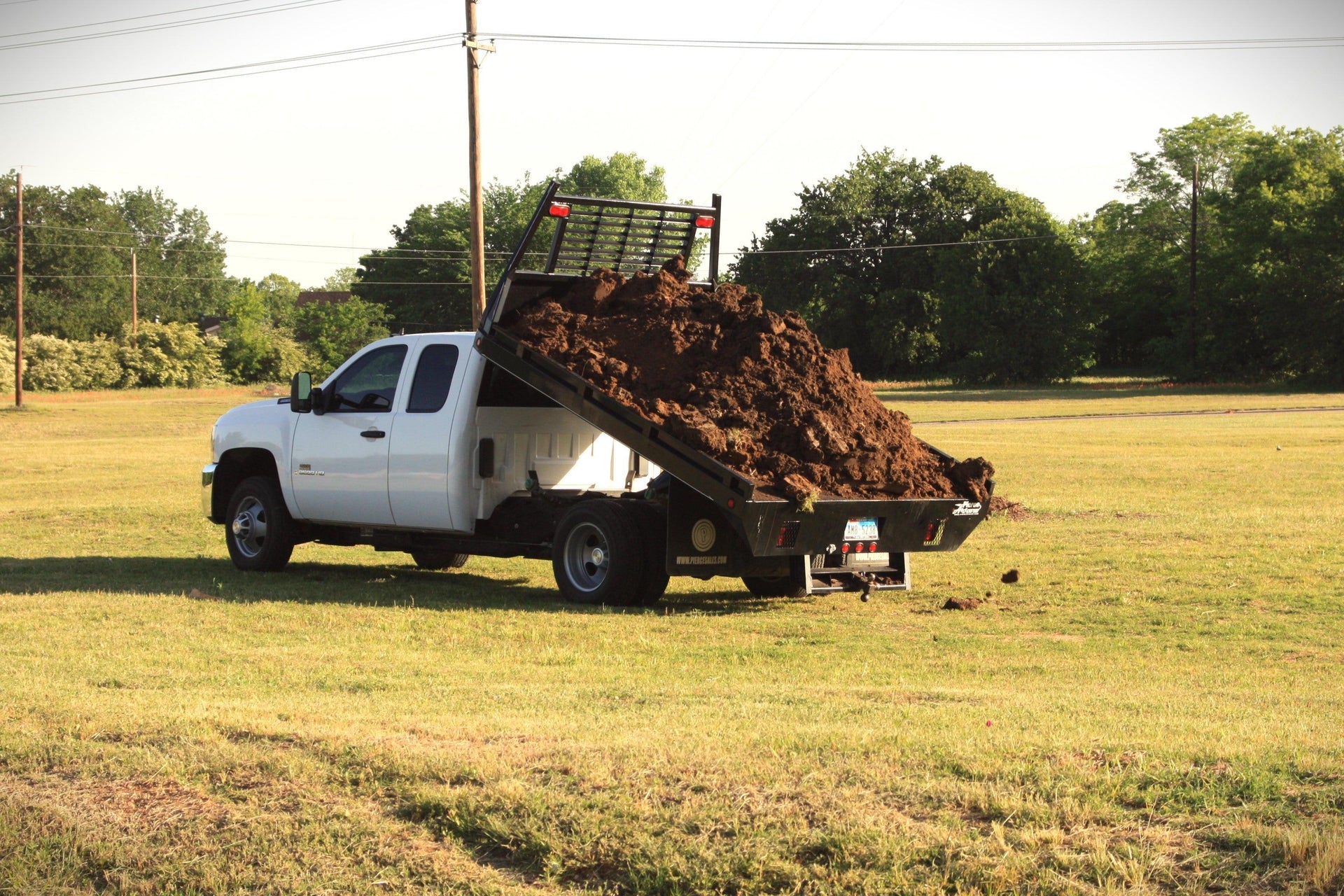A white dump truck unloading a large pile of soil onto a grassy field.