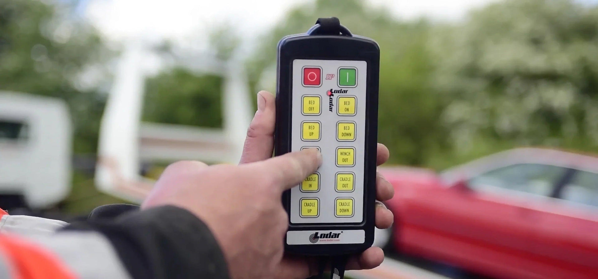 A hand presses a button on a remote control with yellow buttons and a red car in the blurred background.