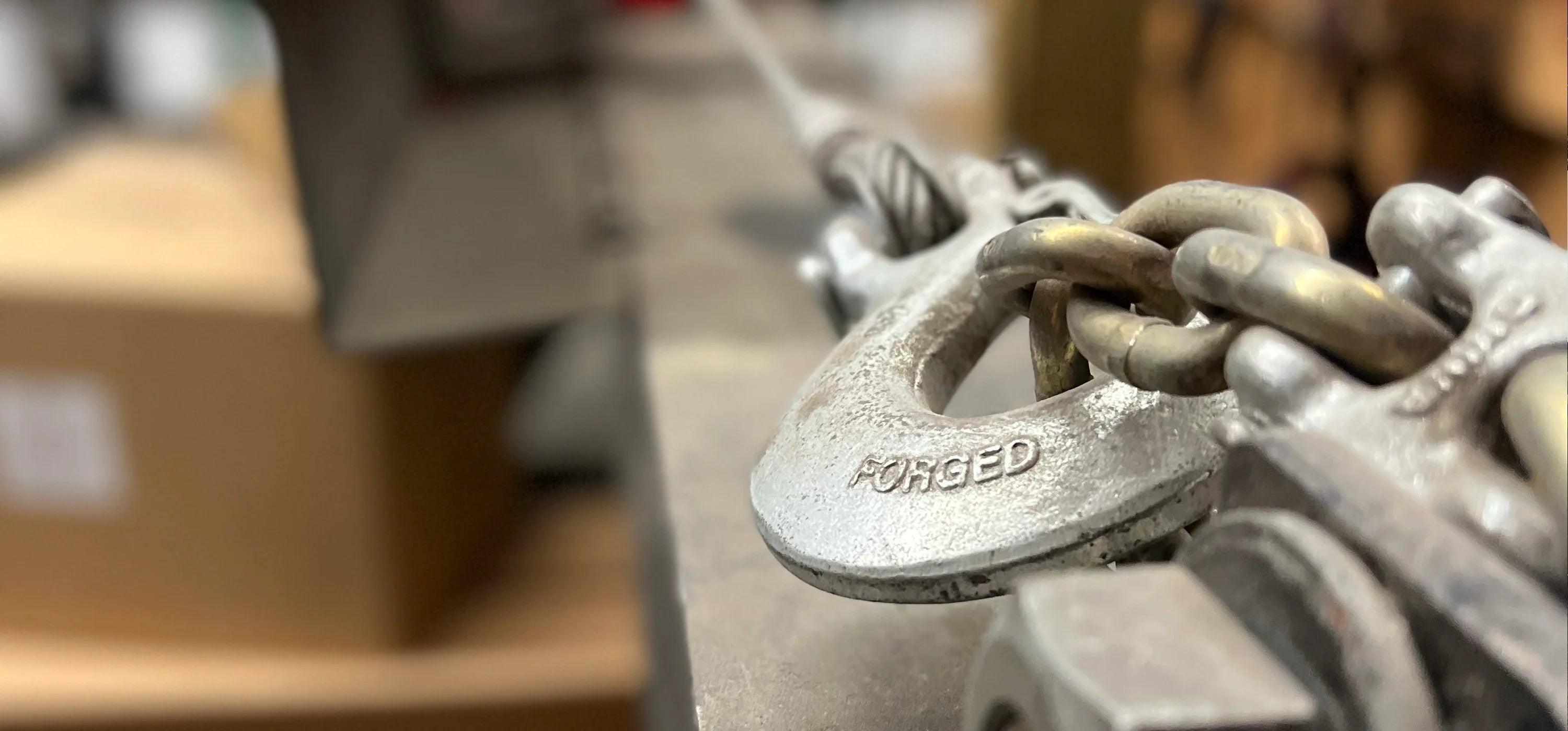Close-up of a metal chain link labeled FORGED, attached to industrial equipment, with a blurred background.