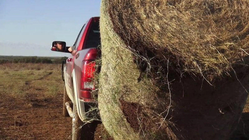A red pickup with a PIERCE Truck Bed Gooseneck Hay Bale Mover hauls a large round hay bale in a rural field as someone’s arm hangs out the window.