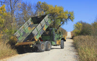 A military dump truck equipped with a PIERCE Heavy Duty Flat Bed Dump Kit is parked on a rural, tree-lined gravel road with its bed raised.