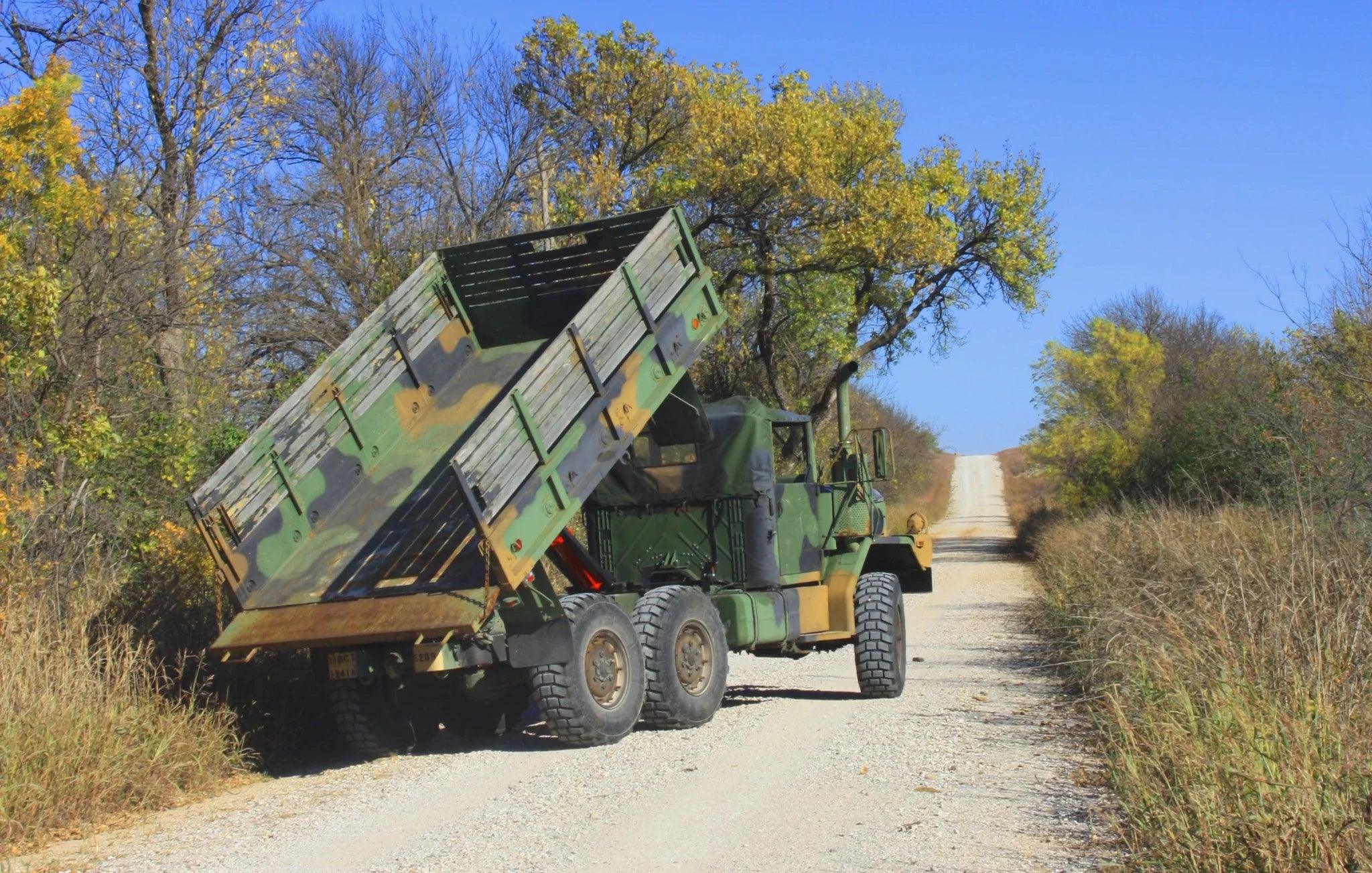A military dump truck equipped with a PIERCE Heavy Duty Flat Bed Dump Kit is parked on a rural, tree-lined gravel road with its bed raised.