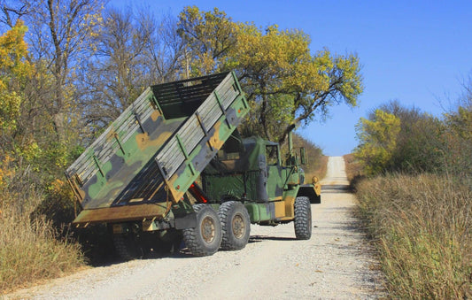 A military dump truck with a PIERCE Heavy Duty Flat Bed Dump Kit, featuring a 20,000-lb. lifting capacity, is parked on a gravel road among trees and grass.