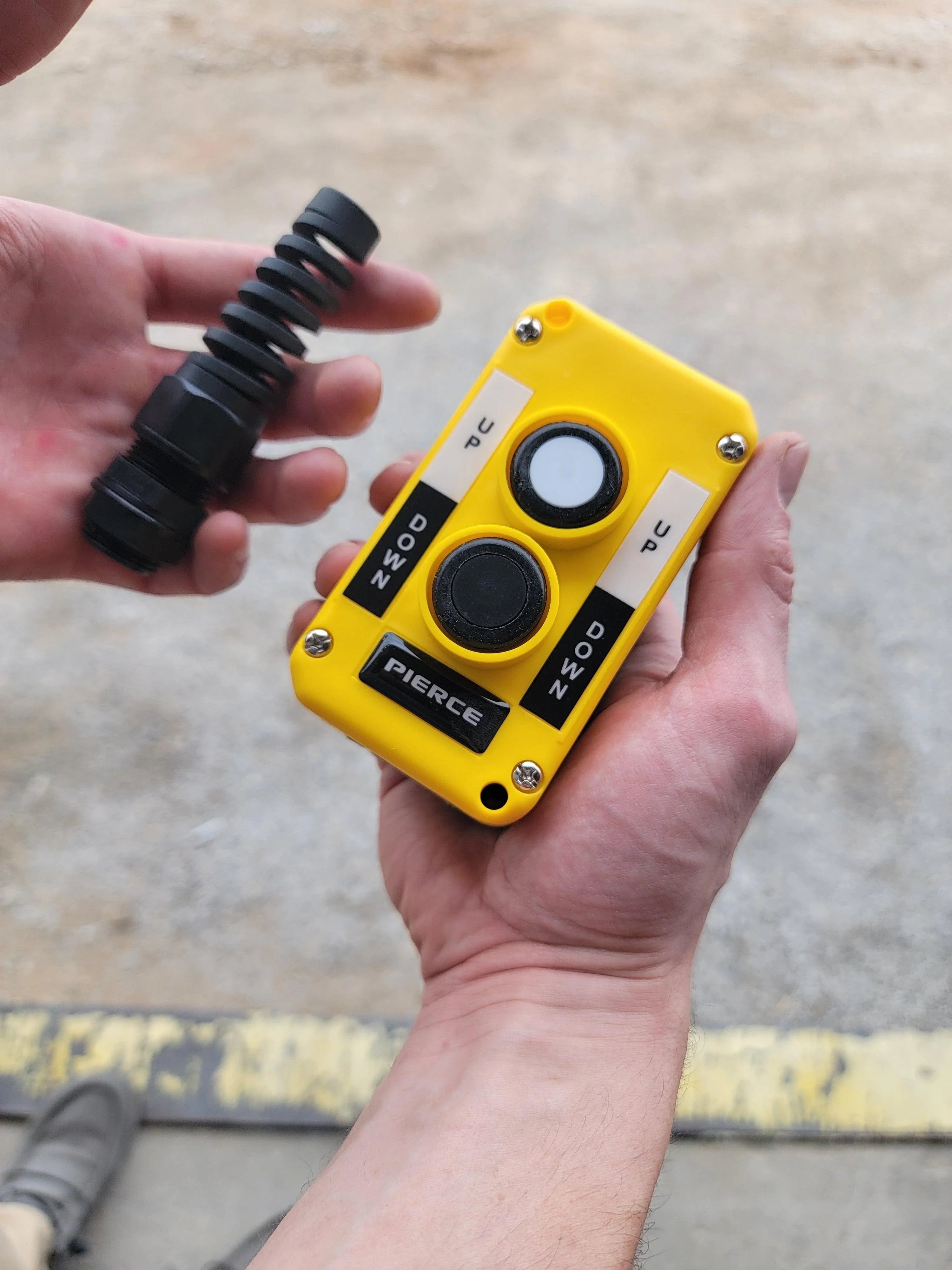 A person holds a yellow Asetek Enterprise Company LTD Universal Momentary Pendant Control - 2 Button and a black rubber part, both essential for hydraulic power units in industrial settings.