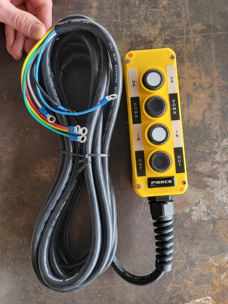 A hand holds a PIERCE Universal Momentary Pendant Control, featuring four buttons and attached colored wires, on a metal surface. This yellow control panel, which includes a 20ft cord and magnets, is ideal for managing solenoid-operated equipment.
