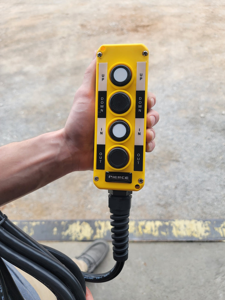 Close-up of a hand holding a PIERCE Universal Momentary Pendant Control with four buttons and a 20-foot cord, featuring magnets. The yellow pendant control offers precise operation for managing solenoid-operated equipment, including buttons labeled Up, Down, and In/Out.