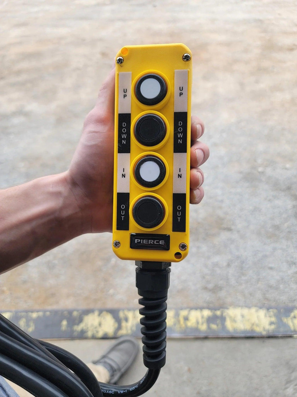 A hand holds the PIERCE Universal Momentary Pendant Control with 4 buttons (Up, Down, In, Out), a 20ft cord, and magnets—ideal for solenoid-operated equipment.
