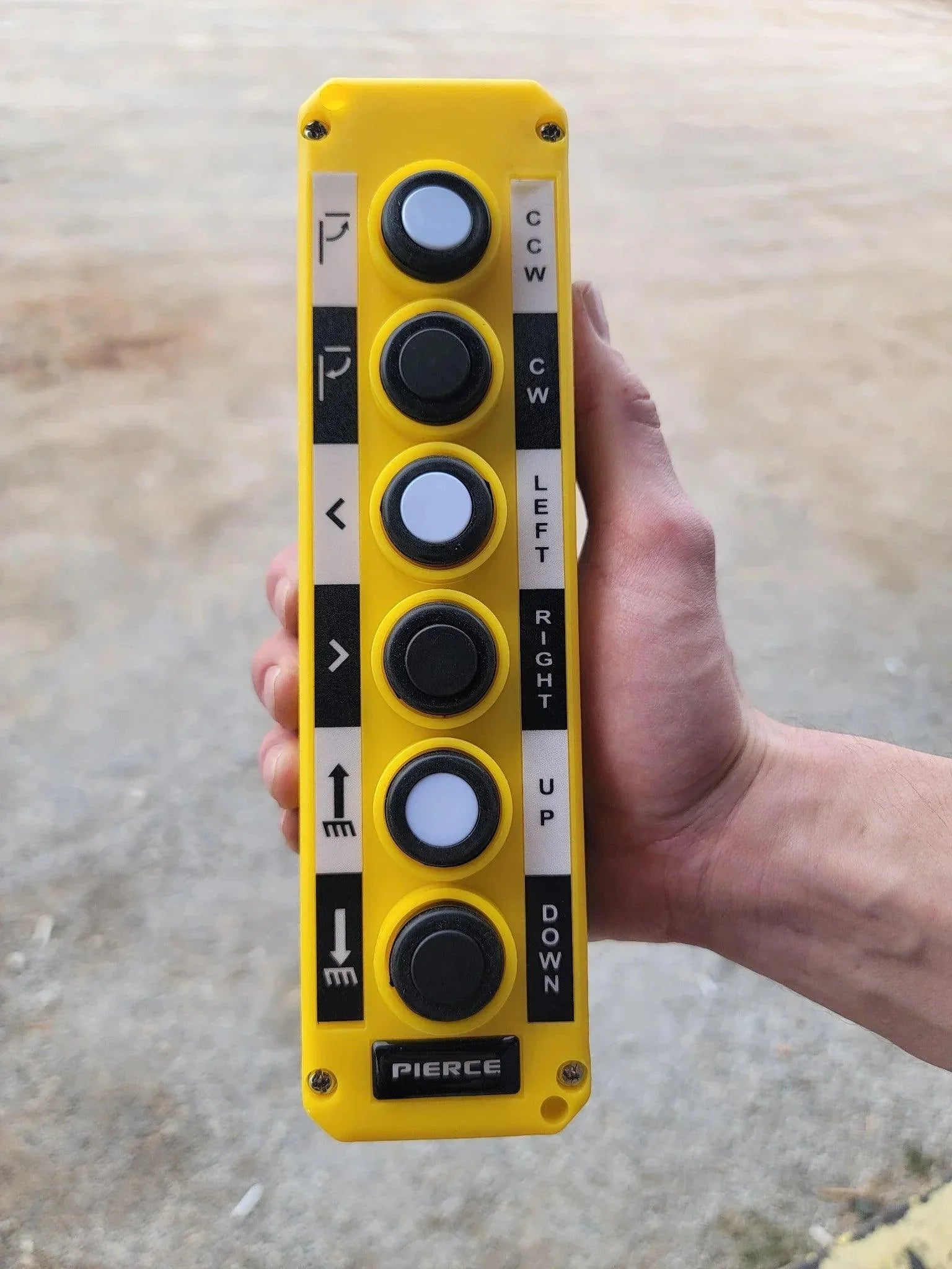 A hand holds an Asetek Enterprise Company LTD Universal Momentary Pendant Control - 6 Button, a yellow device with labeled black buttons for directional movement, commonly used to operate hydraulic power units.