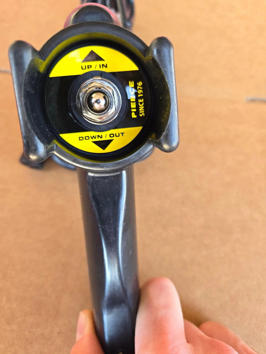 A close-up of the PIERCE Universal Toggle Switch Remote - No Cord in black, with a yellow UP/IN and DOWN/OUT label, resembling a replacement pendant by PIERCE, set against a brown background.