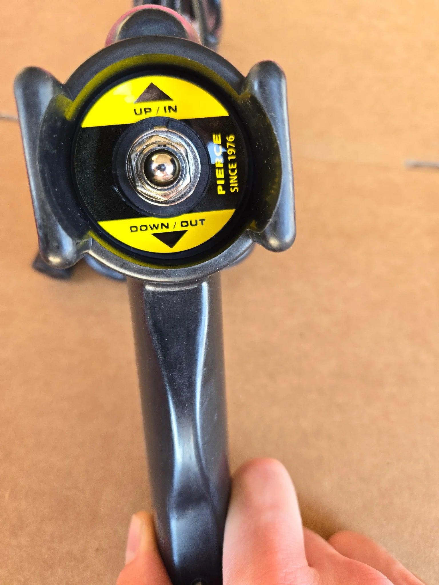 A close-up of the PIERCE Universal Toggle Switch Remote - No Cord in black, with a yellow UP/IN and DOWN/OUT label, resembling a replacement pendant by PIERCE, set against a brown background.