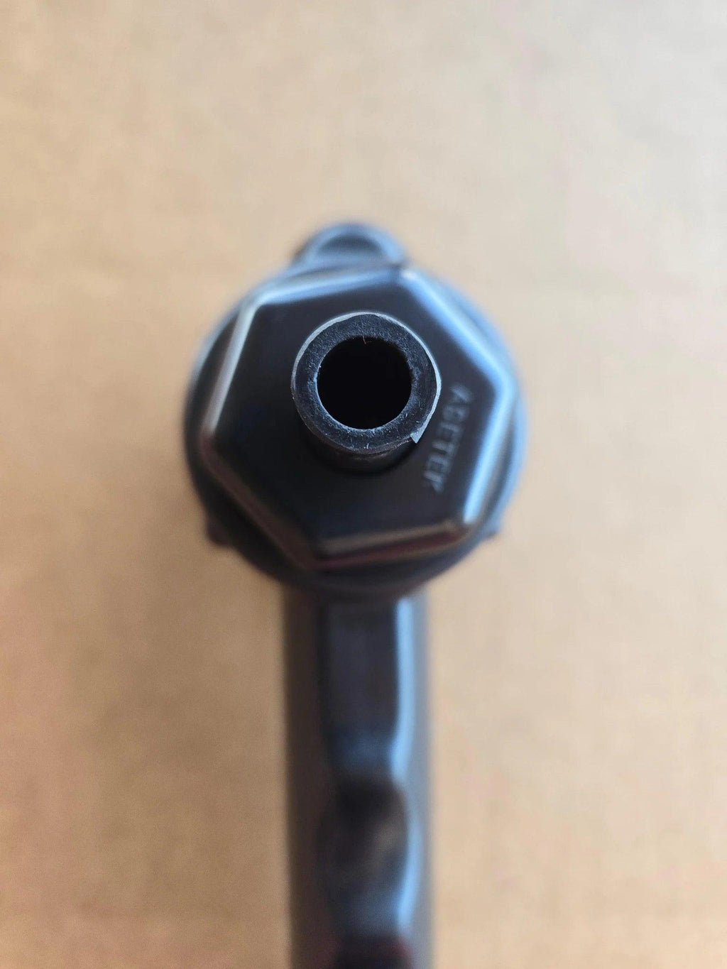 Close-up of the PIERCE Universal Toggle Switch Remote - No Cord by PIERCE, a black hexagonal device with a central hole, viewed from above on a tan background.