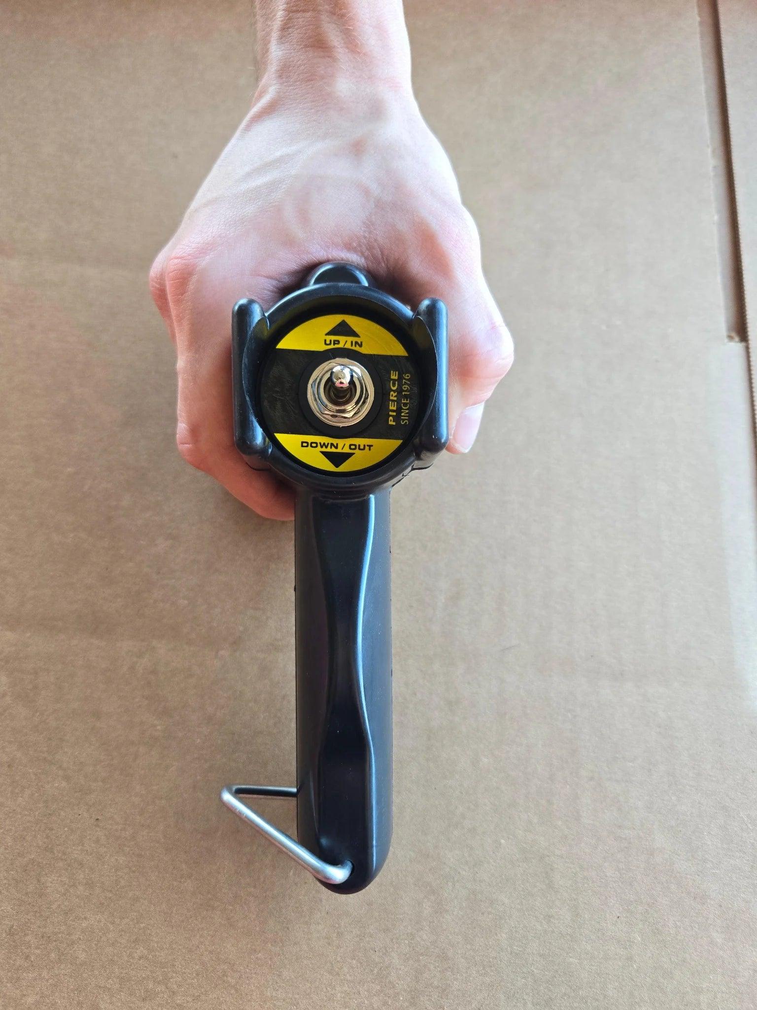 A hand holds a black PIERCE Universal Toggle Switch Remote - No Cord with a yellow circular label on a brown cardboard surface.