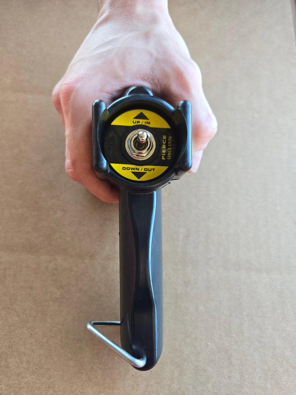 A hand holds a black and yellow PIERCE Universal Toggle Switch Remote - No Cord, labeled UP/IN and DOWN/OUT, against a brown background.