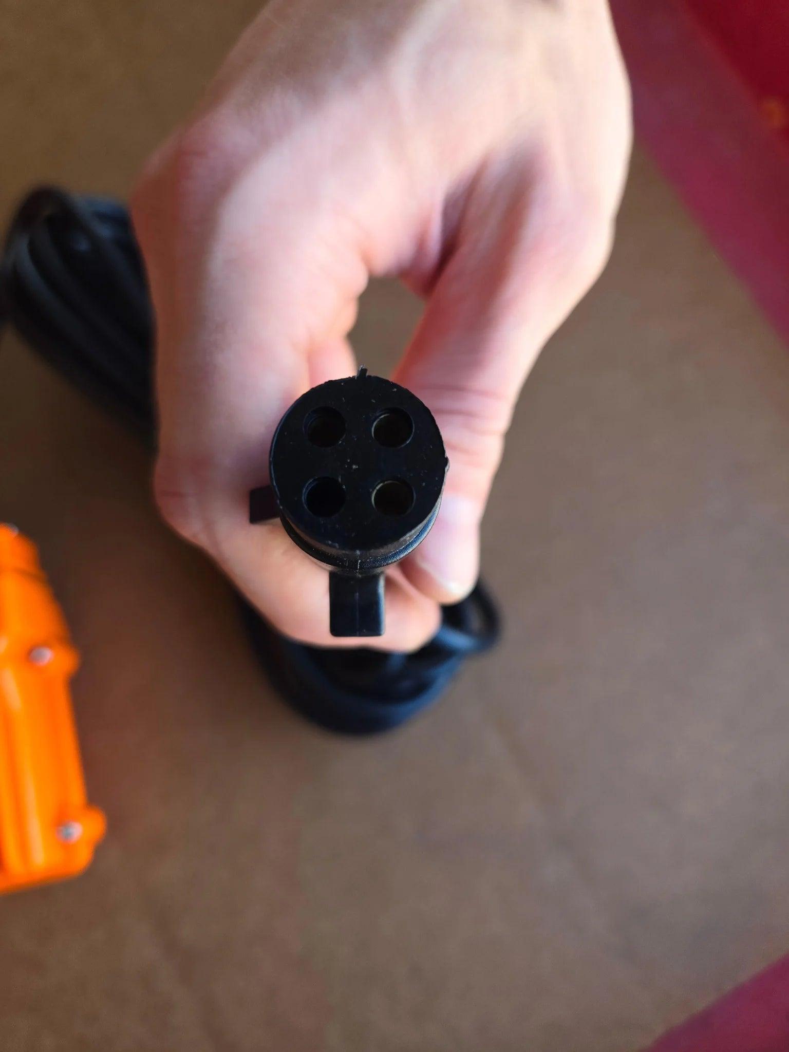 A hand holds the black 4-prong vulcanized plug of the PIERCE 2 Function Momentary Hard-Wired Control Pendant (15 ft. cord) by Asetek Enterprise Company LTD, with a blurred orange control visible nearby.