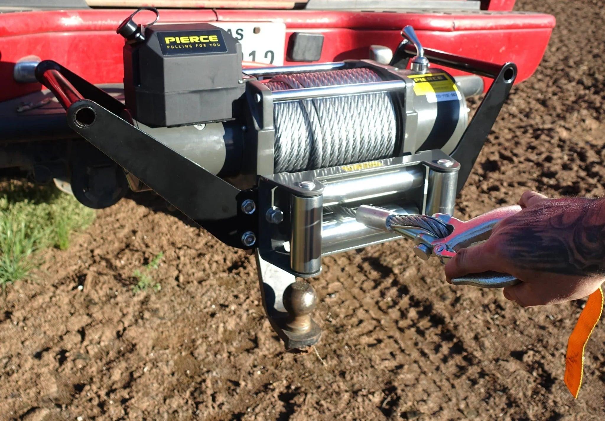 A hand attaches a PIERCE 20,000 lb Recovery Winch cable to the rear of a red truck parked on dirt.
