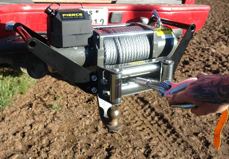A hand attaches a PIERCE 20,000 lb Recovery Winch cable to the rear of a red truck parked on dirt.