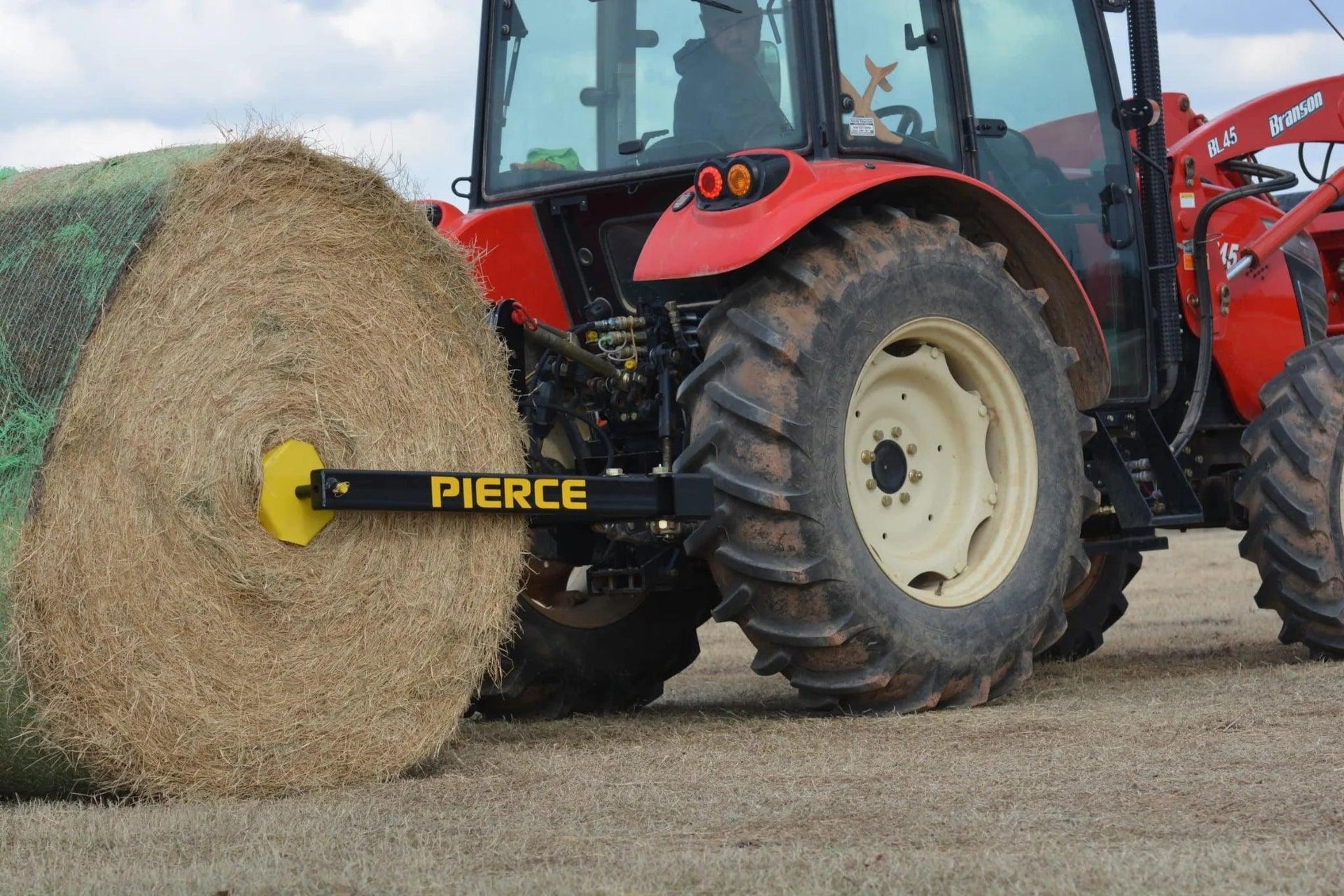 A red tractor equipped with the Internal - Pierce Assembly Team 3-Point Hay Bale Unroller moves a large round hay bale across the field.