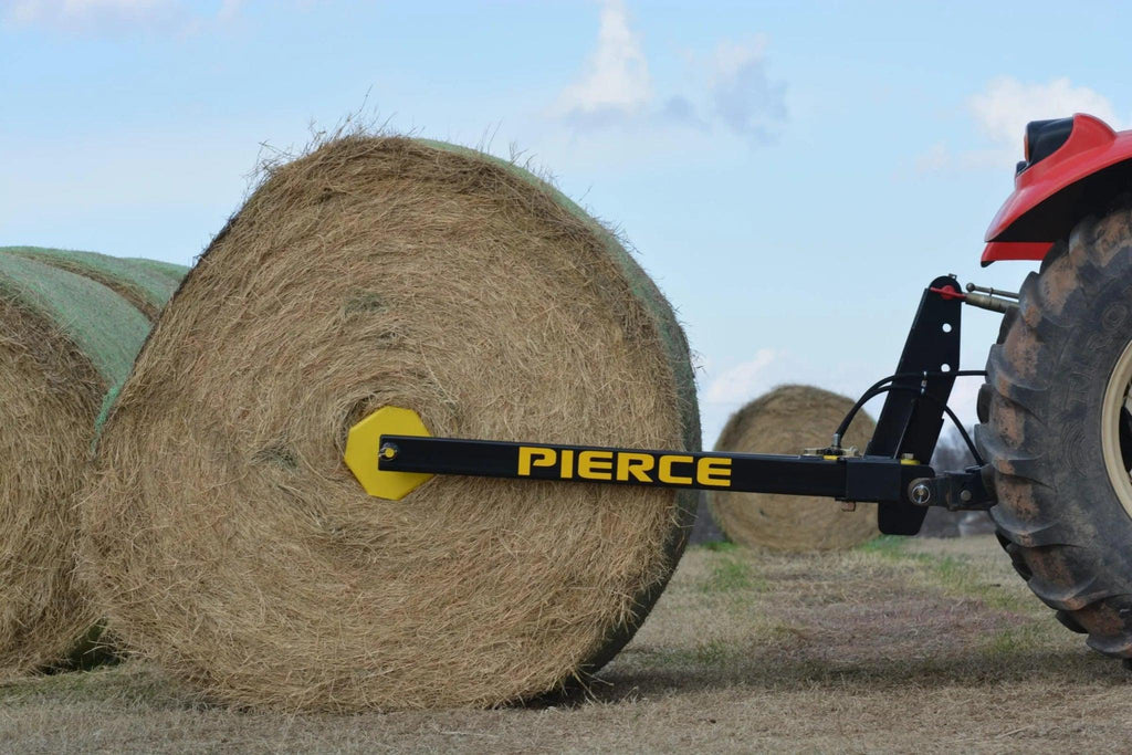 A tractor lifts a large hay bale in a field using an Internal - Pierce Assembly Team 3-Point Hay Bale Unroller attachment.