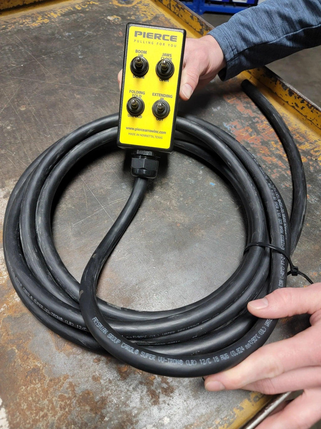 A person holds a PIERCE 8 Function Wrecker Remote, featuring buttons and a long coiled black cable on a metal surface—perfect for operating 12/24V solenoid systems or hydraulic power units.