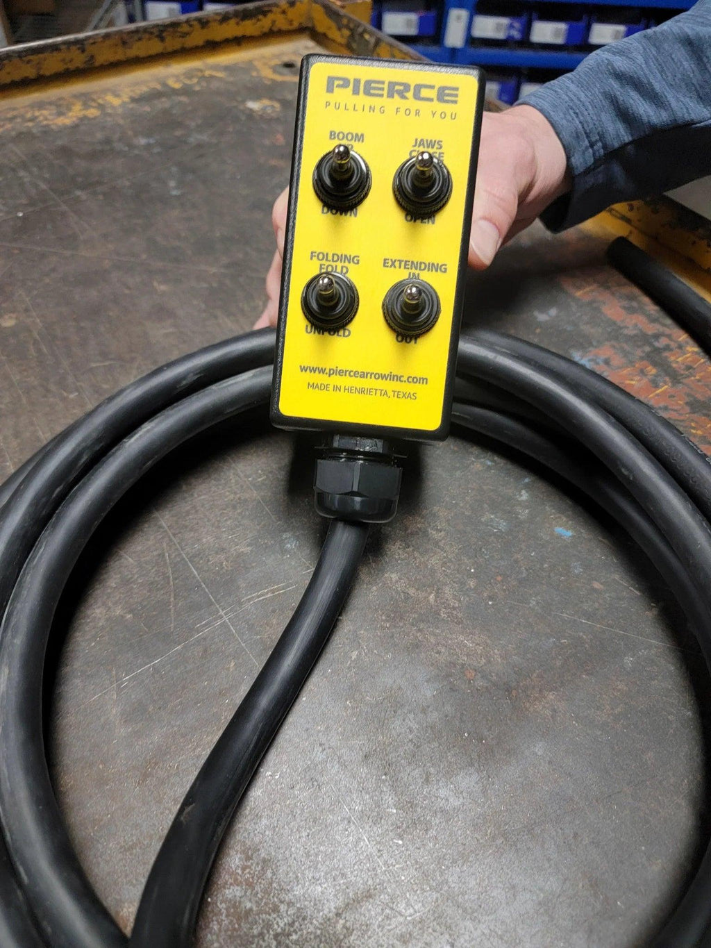 A hand holds a yellow PIERCE 8 Function Wrecker Remote with four labeled switches and a thick black cable, designed for 12/24V solenoid systems, resting on a metal surface.