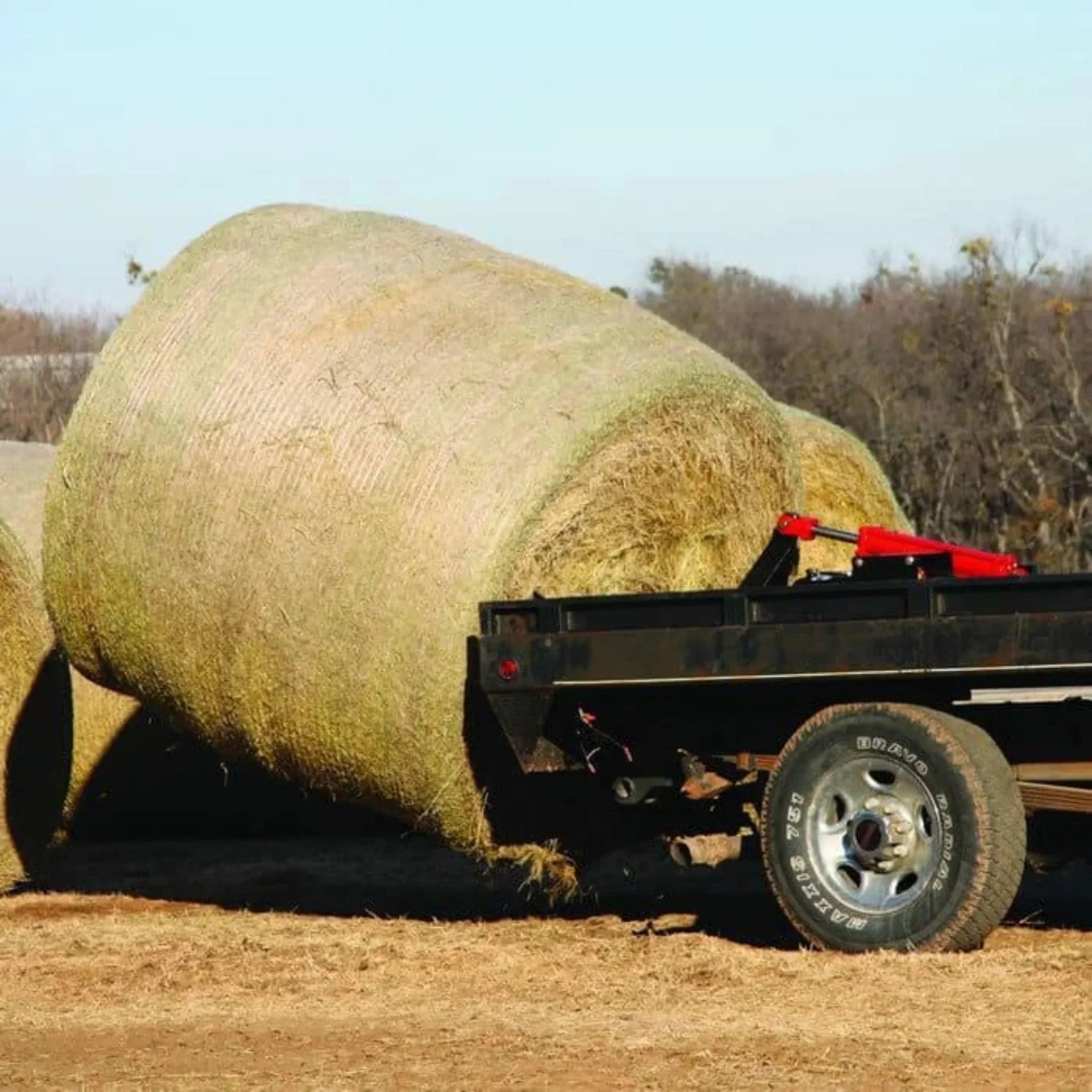 A large hay bale rests on a flatbed trailer in a rural setting, with trees behind and the PIERCE Bolt-in Hydraulic Truck Bed Hay Bale Spear ready for easy loading.