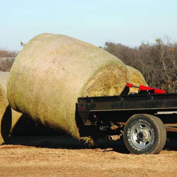 A large hay bale rests on a flatbed trailer in a rural setting, with trees behind and the PIERCE Bolt-in Hydraulic Truck Bed Hay Bale Spear ready for easy loading.