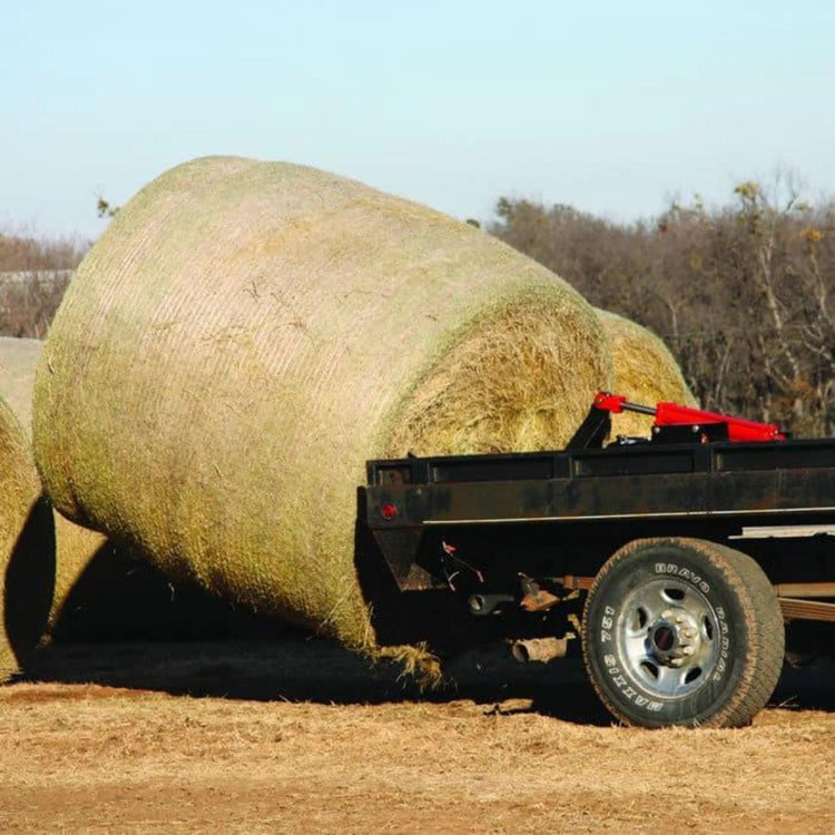 Large round hay bales rest beside a flatbed trailer with trees behind, ready to be loaded using the PIERCE Truck Bed Gooseneck Hay Bale Mover.