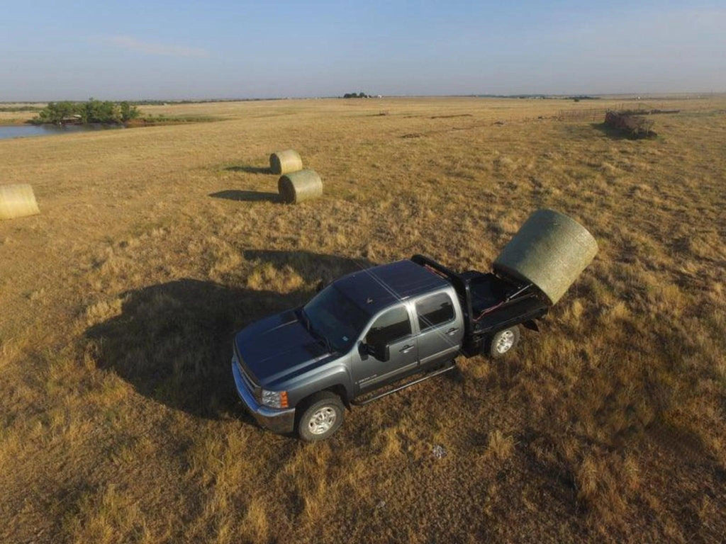 A PIERCE Bolt-in Hydraulic Truck Bed Hay Bale Spear unloads a large round hay bale in a dry field, with more bales scattered in the background.