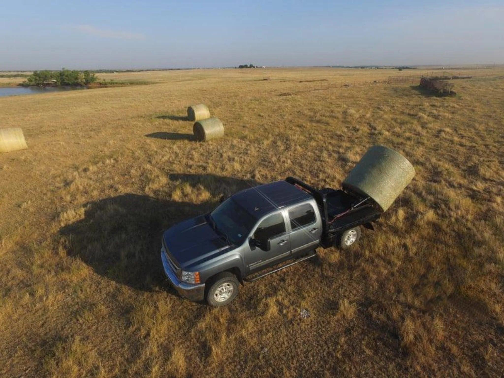 A black pickup truck equipped with a PIERCE Bolt-in Hydraulic Truck Bed Hay Bale Spear unloads a large hay bale in a field with more hay bales scattered nearby.