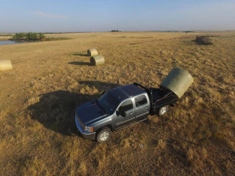 A black pickup truck equipped with a PIERCE Bolt-in Hydraulic Truck Bed Hay Bale Spear unloads a large hay bale in a field with more hay bales scattered nearby.