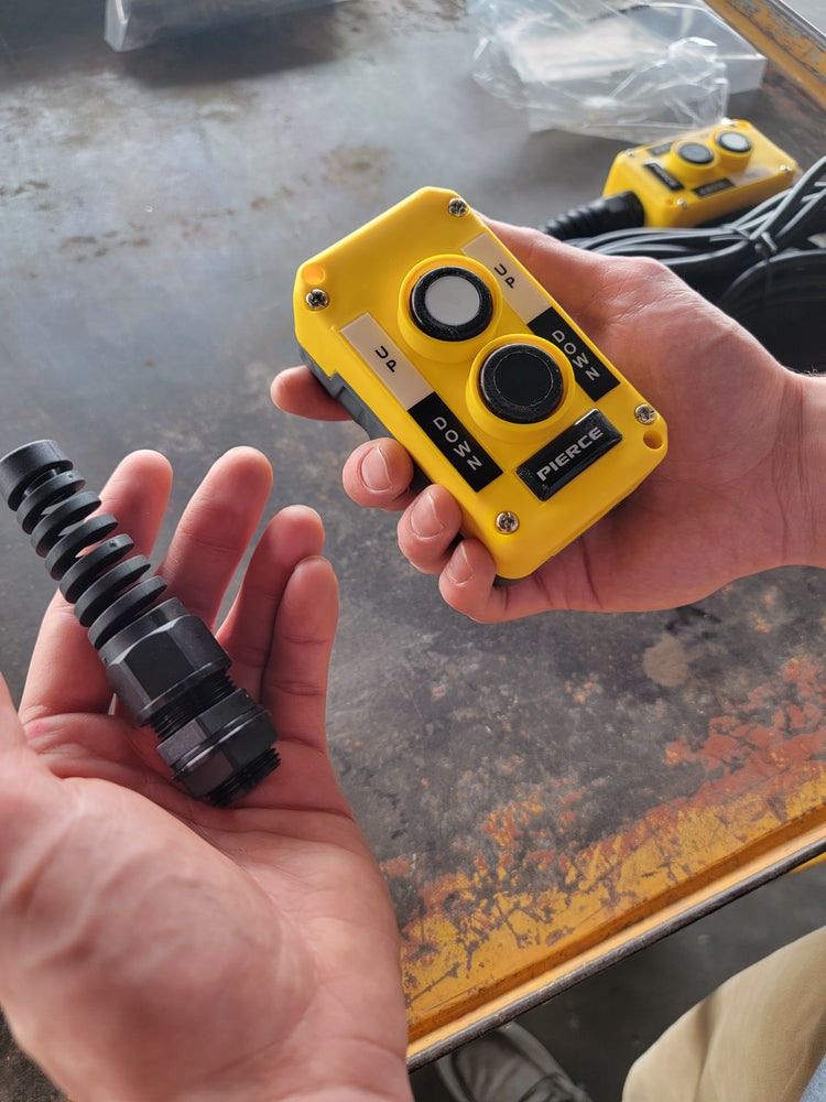 Hands holding a PIERCE Universal Momentary Pendant Control - 2 Button and a black cylindrical component, both used in industrial settings, rest on the table.