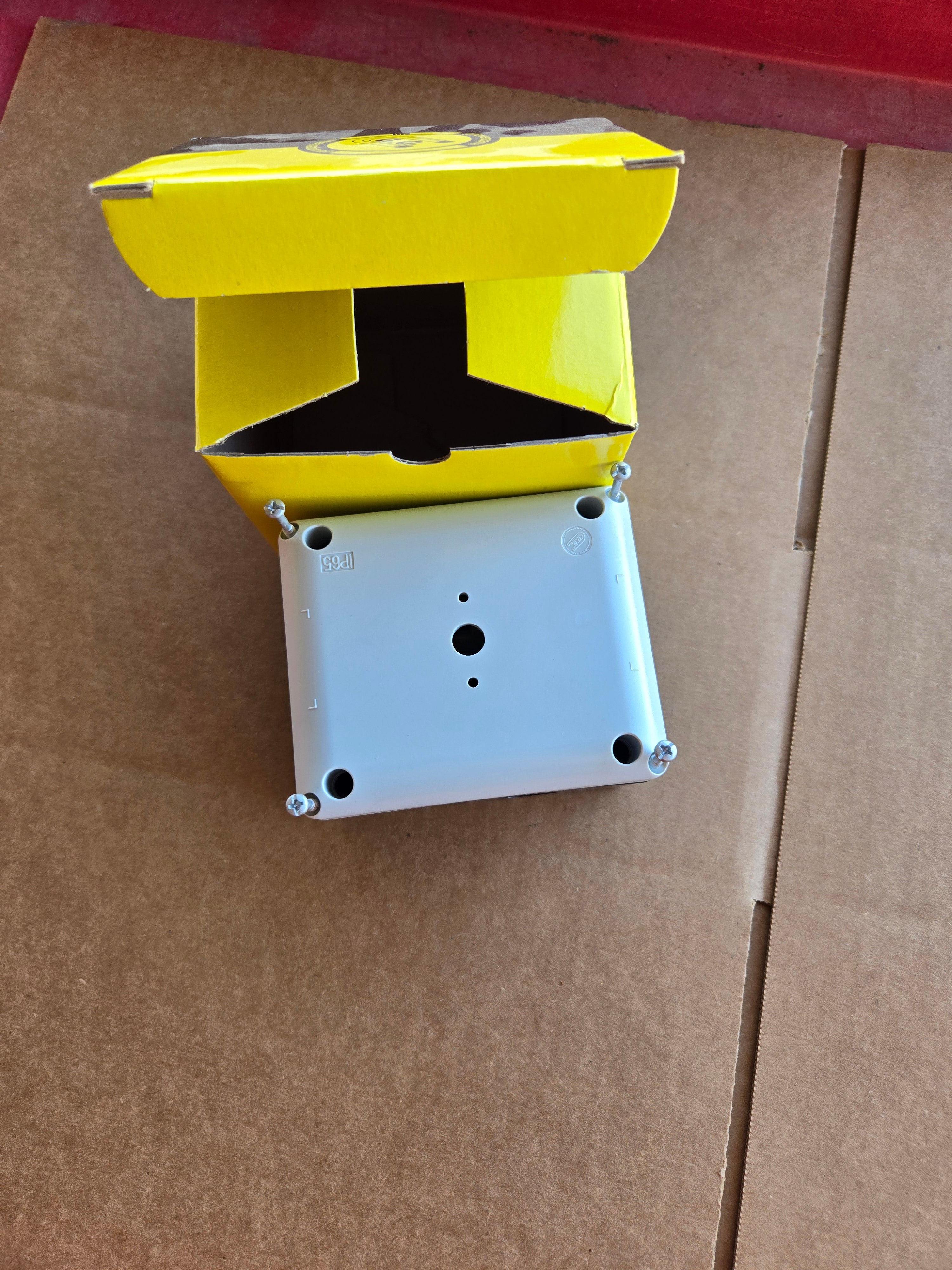 A gray junction box, resembling a switch enclosure, peeks from a yellow box on cardboard. Perfect for industrial use, its the PIERCE Control Box for 110V PIERCE 654 Series Single Phase Winch, ideal for efficiently managing single-phase motors.