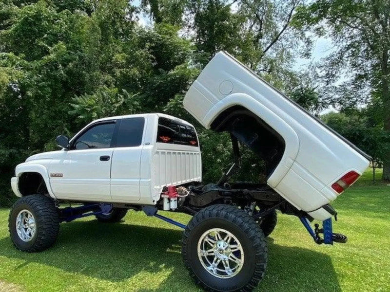 A white lifted pickup truck with a PIERCE Factory Bed Dump Kit has its bed raised vertically, shown on green grass with trees behind.