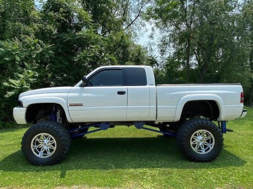 White lifted pickup truck with large off-road tires and a PIERCE Factory Bed Dump Kit installed, parked on grass with trees in the background.