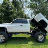 A white lifted pickup truck equipped with the PIERCE Factory Bed Dump Kit has its bed tilted up, parked on grass with trees in the background.