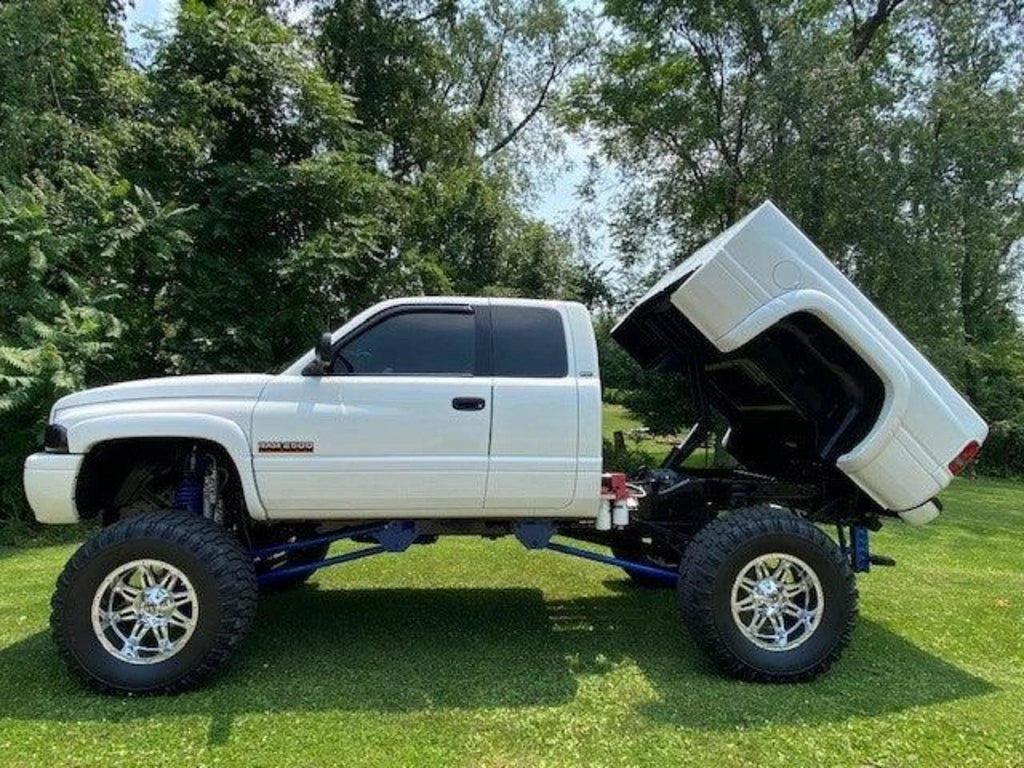 A white lifted pickup truck equipped with the PIERCE Factory Bed Dump Kit has its bed tilted up, parked on grass with trees in the background.